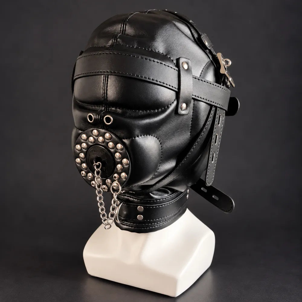Black leather bondage hood with covered eyes, adjustable leather straps, silver studs, circular mouth cover and metal chains, displayed on a white mannequin in three-quarter front view.
