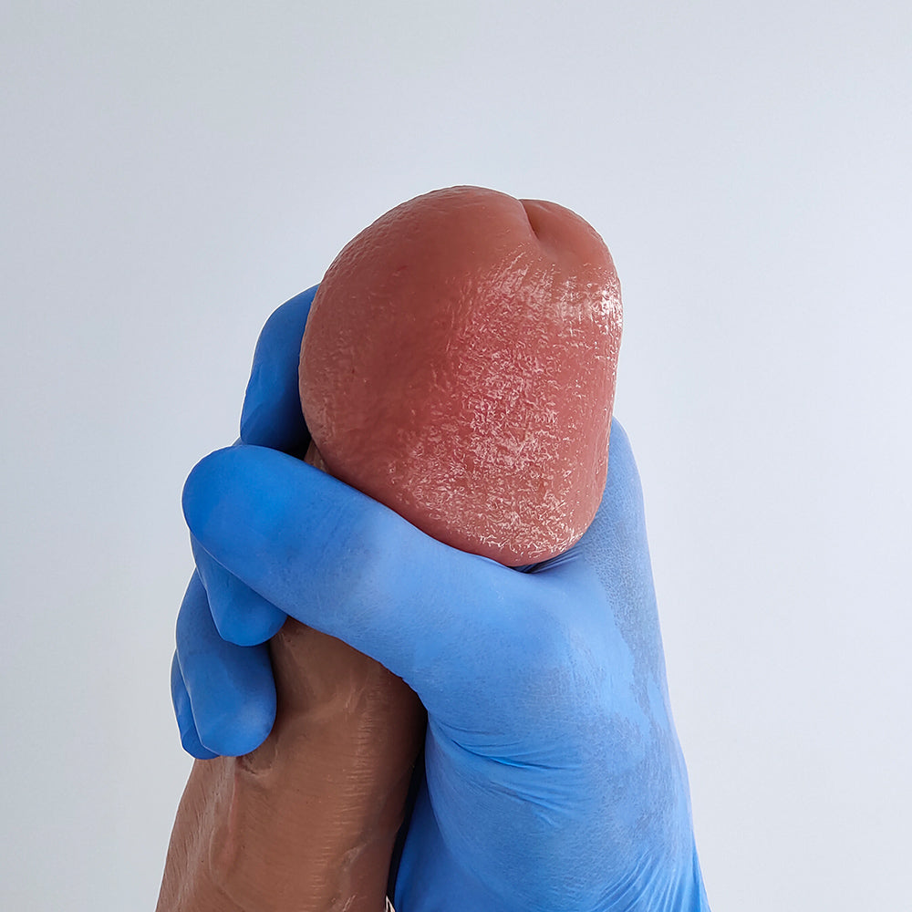 Close-up of the glans on the Classic penis sleeve held in hand with a blue glove, showing realistic texture and pigmentation on a light background.
