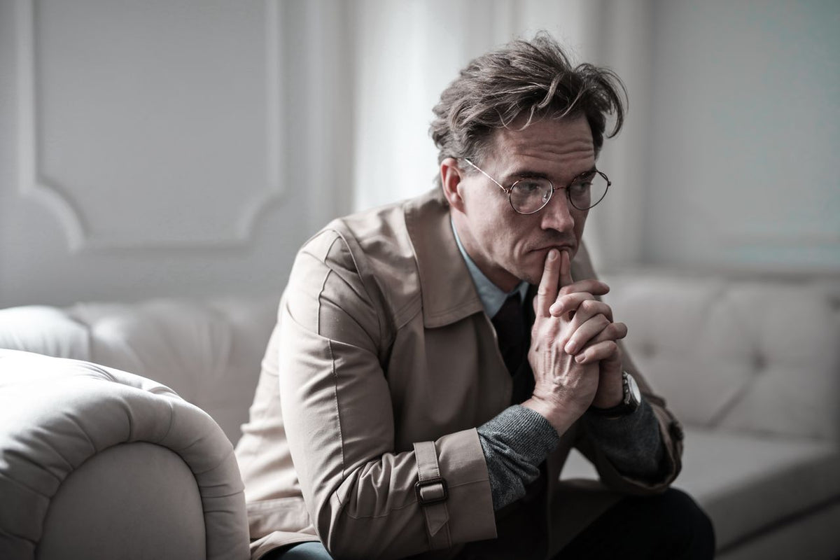 A thoughtful businessman with round glasses and a beige trench coat, sitting on a white tufted leather sofa with his hands clasped to his face.