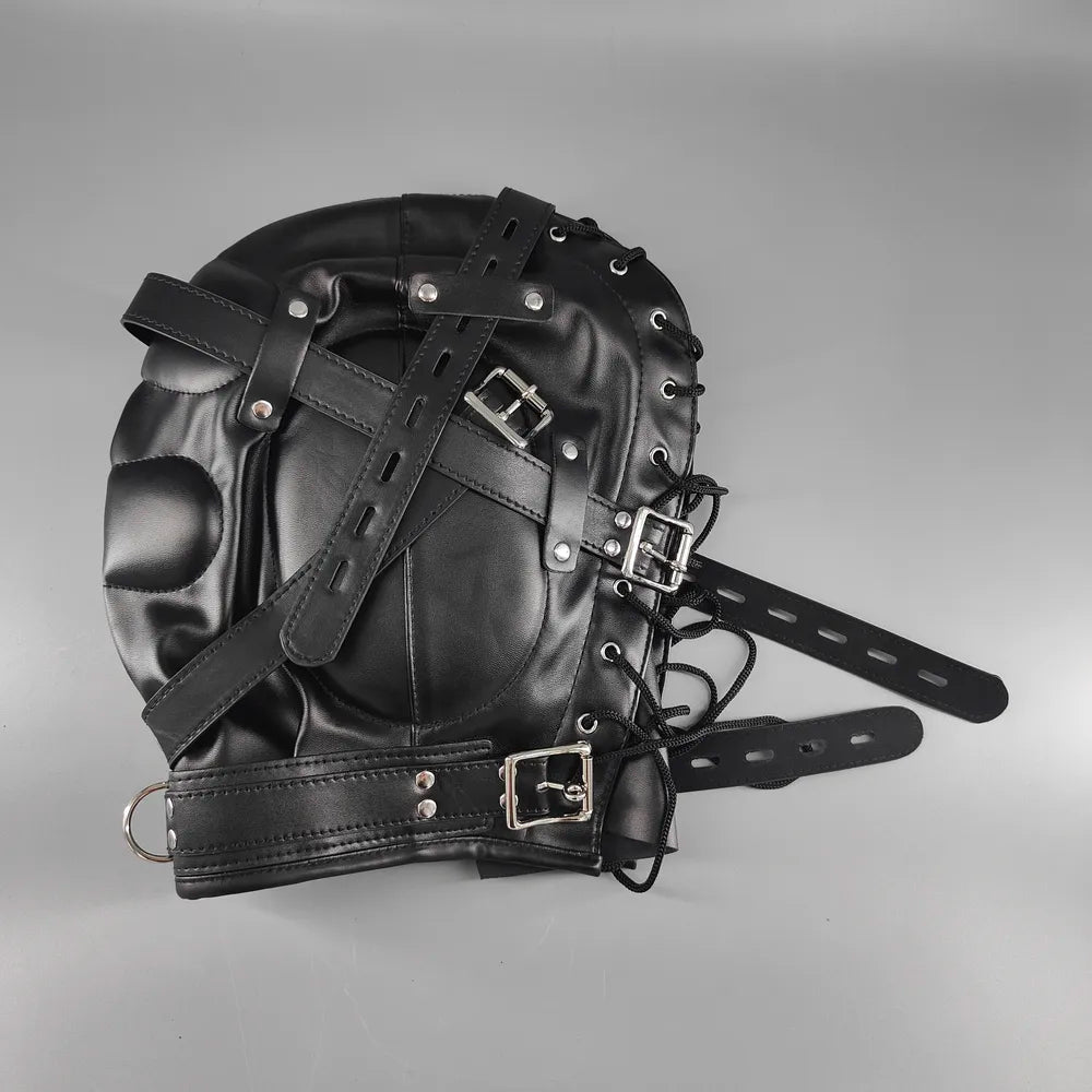 Black leather bondage hood lying on its side on a gray surface without a mannequin, showing its padded construction, adjustable straps, and metal hardware.