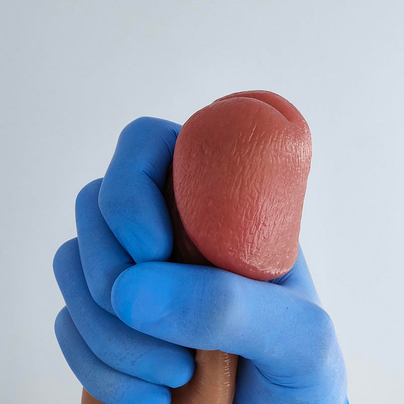 Side view close-up of the glans on the Classic penis sleeve, held in hand with a blue glove, showing curved shape and realistic silicone texture on a light background.
