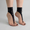 Side view of a minimalist latex foot harness with stainless steel D-ring and toe strap worn on tiptoe against a light gray studio background